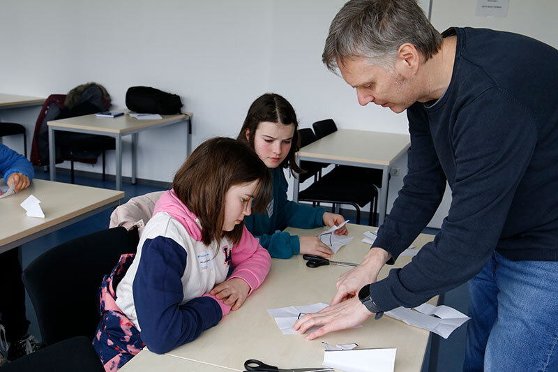 Prof. André Schulz erklärt zwei Mädchen, wie sie ein Blatt falten sollen.