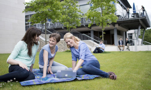 Drei Studentinnen sitzen auf der Wiese vor der Mensa mit einem aufgeklappten Laptop auf der Decke.