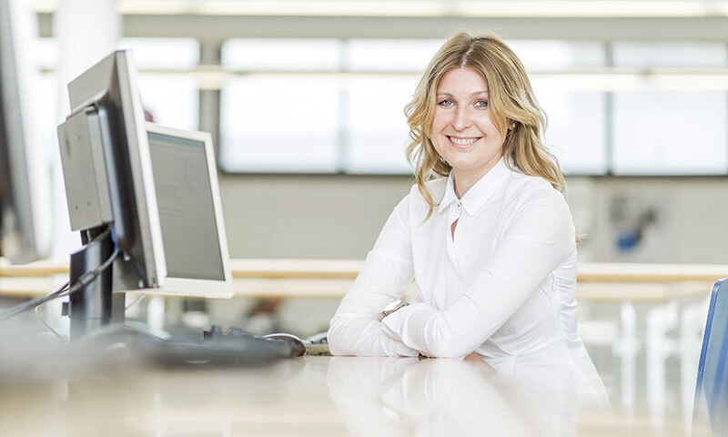 Blonde Frau am Bürotisch mit zwei Bildschirmen in heller Umgebung dem Betrachter lächelnd zugewandt.