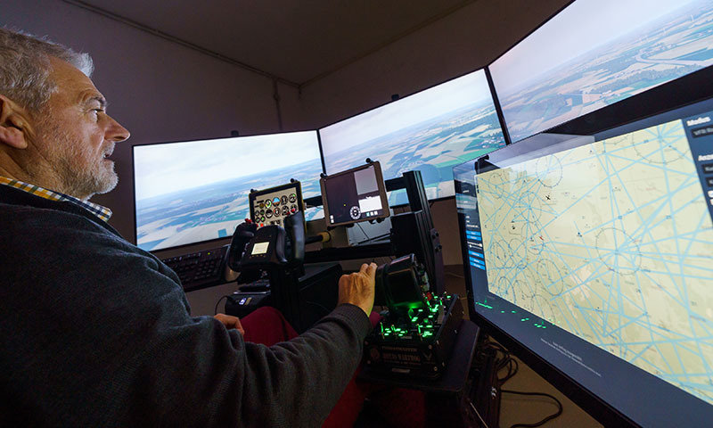 Ein Mann sitzt vor Gerätschaften, die ein Flugzeugcockpit simulieren, und blickt auf mehrere Computerbildschirme, auf denen aus der Pilotenperspektive Landschaft zu sehen ist.