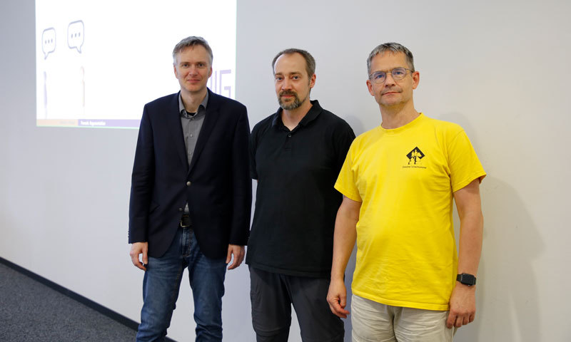 Drei Männer stehen vor einer Leinwand. Die Präsentation wird angezeigt.