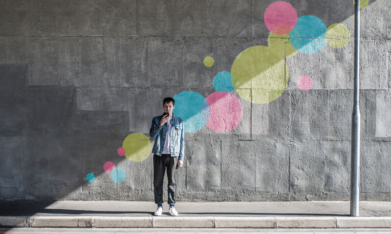 Mann, auf sein Smartphone schauend, steht vor einer grauen Wand an einer Straße. Auf der Wand befinden sich verschieden große Punkte in gelb, rosa und blau. Links neben dem befindet sich eine Straßenlaterne.