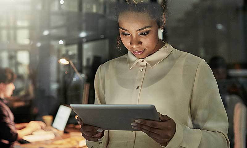 Junge Frau im  Büro mit einem Tablet in der Hand. Die Frau bildet sich im Compliance weiter. Sie nutz dazu den Selbstlernkurs „Compliance“ der Fernuniversität. Im Hintergrund sitzt ein Mann an einem Computer und arbeitet.