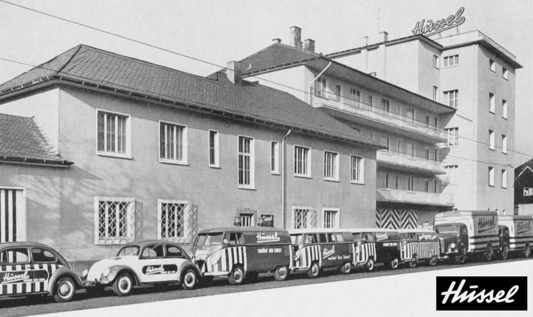 Schwarz-weiß-Fotografie der Hussel Firmenzentrale aus dem Jahr 1957 von der gegenüberliegenden Straßenseite aus fotografiert. Vor dem Gebäude stehen VW-Käfer und VW-Bullis, mit dem Firmenlogo versehen, in einer Reihe geparkt.