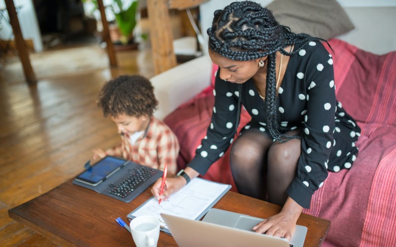 Eine Schwarze Frau sitzt mit ihrem Kind auf dem Sofa, sie macht sich Notizen, vor ihr steht ein Laptop, das Kind tippt auf einem Tablet.