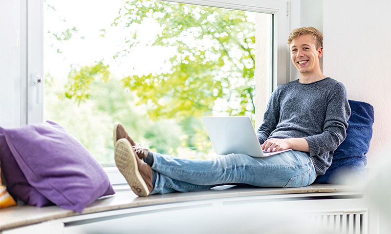 Studierender mit Laptop entspannt auf einer Fensterbank