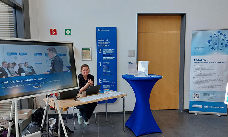 Frau sitzt am Info-Tisch zwischen Videoscreen, Stehtisch und Rollup