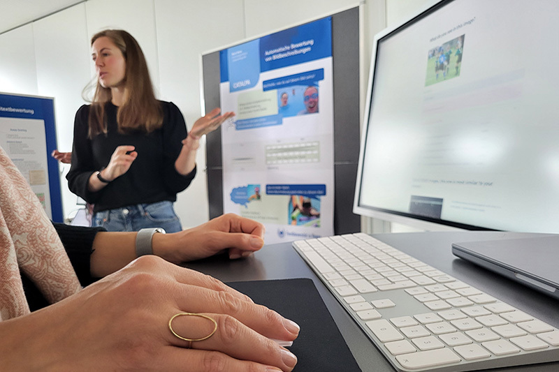 Im Vordergrund ein Monitor und zwei Hände an einer Tastatur, im Hintergrund eine junge Frau, die etwas erklärt
