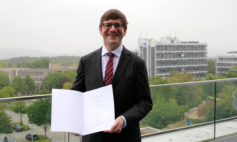 Professor Kresse smiling and standing on a balcony overlooking the university, holding a certificate