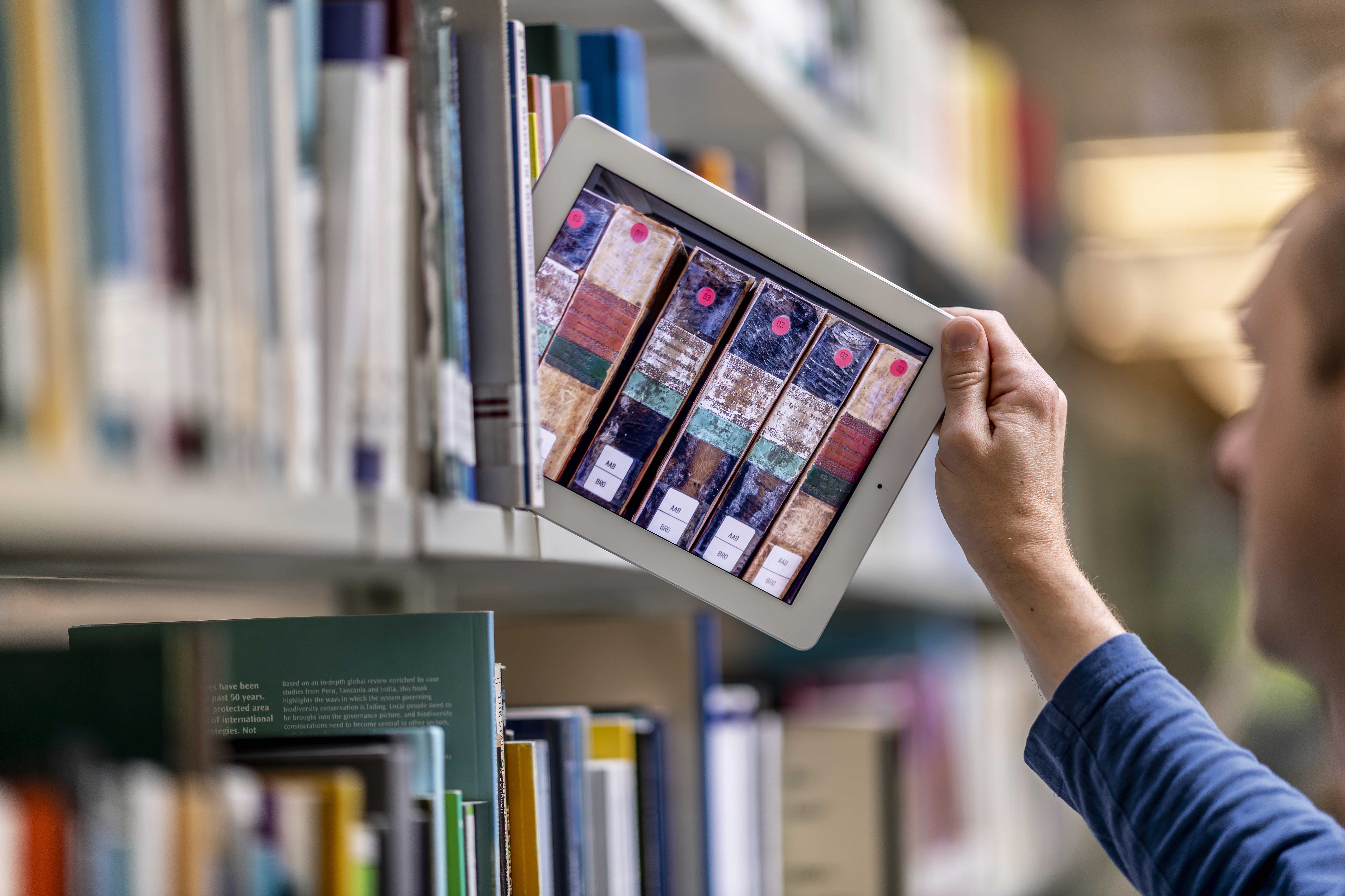 Es ist ein Bücherregal zu sehen aus der eine Person ein Tablet zieht auf dem digital weitere Buchrücken zu sehen sind.