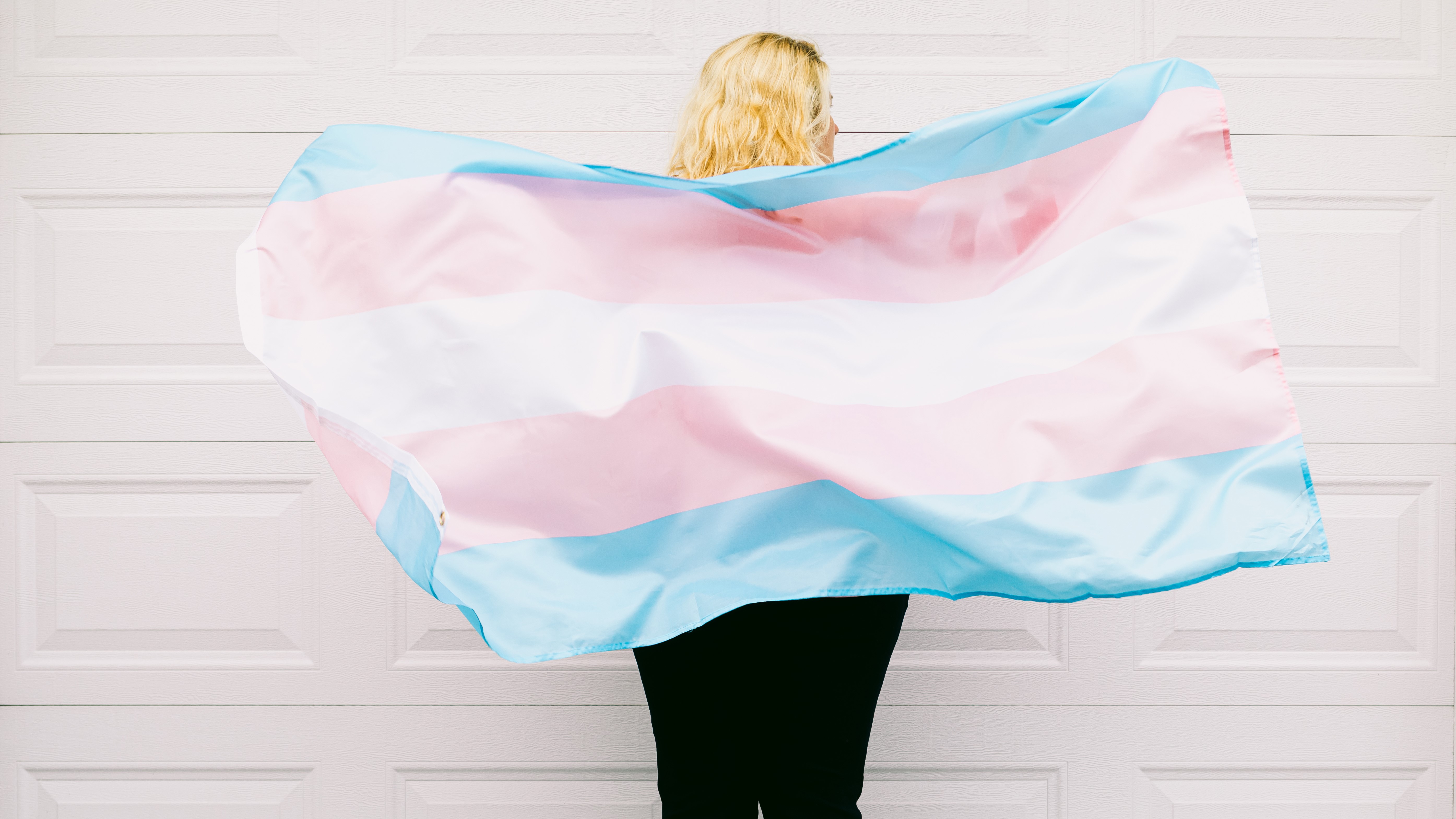 Weiße Wand, davor steht eine Person mit längeren Haaren mit dem Gesicht zur Wand. Die Person hält vor ihrem Rücken die Transgender-Flagge.