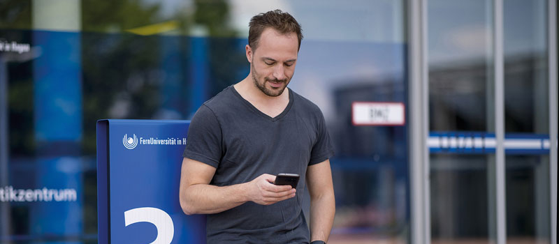 junger Mann lehnt mit Smartphone an FernUni-Stele
