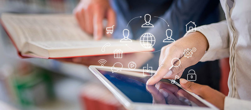 Foto mit Buch und Tablet auf denen mit Fingern etwas gezeigt wird darüber sind grafische Icons zu Onlinethemen