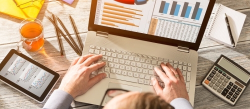 Detailaufnahme aus der Vogelperspektive: Zwei männliche Hände bedienen ein Laptop. Auf dem Dispaly sind ein Torten- und ein Balkendiagramm zu erkennen. Links und rechts davon auf dem Schreibtisch liegen ein Tablet mit weiteren Statistiken und ein Taschenrechner mit Notizblock und Bleistift. Weitere Erkennungsmerkmale: Glas Tee, Brille, weiterer gelber Notizblock und zusätzliche Bleistifte.