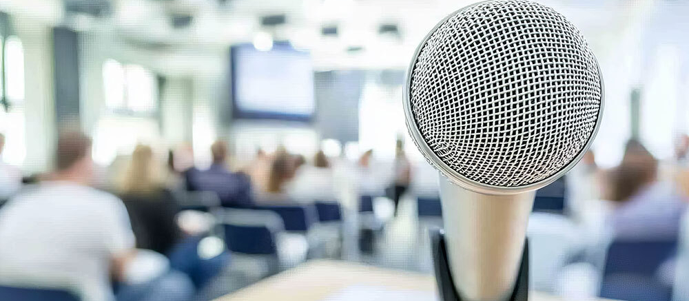 Nahaufnahme eines Mikrofons bei einer Konferenz, mit unscharfem Publikum im Hintergrund