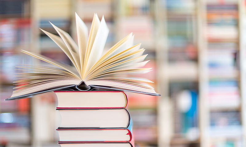 Aufgeschlagenes Buch mit aufgestellten Seiten auf einem Stapel mehrerer geschlossener Bücher vor unscharfem Bücherregal