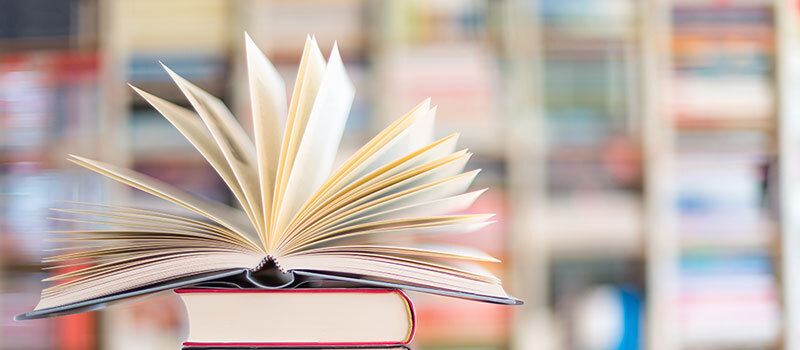 Aufgeschlagenes Buch mit aufgestellten Seiten auf einem Stapel mehrerer geschlossener Bücher vor unscharfem Bücherregal