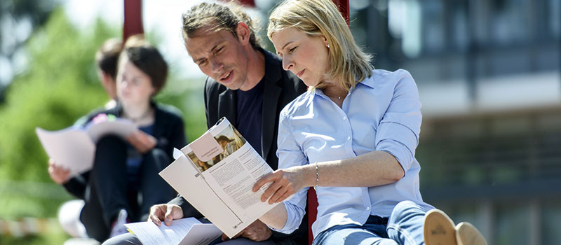 Drei Studentinnen und ein Student sitzen im Vorlesungsraum und schauen gemeinsam in ein aufgeschlagenes Buch, das die Studentin in der Mitte, in ihren Händen hält.