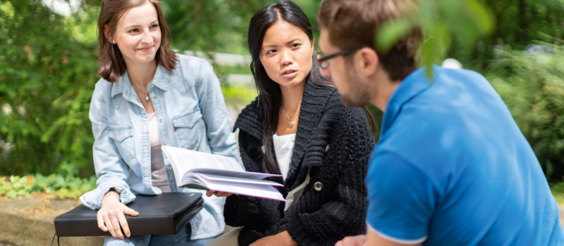 Drei Personen sitzen auf einer Sitzbank im Grünen und unterhalten sich mit ihren Studienunterlagen über die Studieninhalte.
