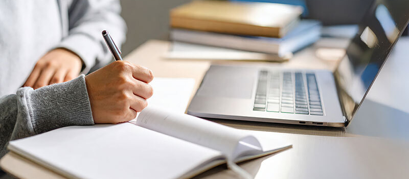 Lernen zu Hause: Hand einer Person, die mit einem Stift in ein aufgeschlagenes Notizbuch schreibt, daneben ein aufgeklappter Laptop auf einem Tisch.