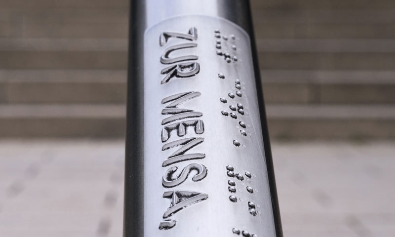 Ein silbernes Treppengeländer wird mit der Aufschrift „Zur Mensa“ in Schwarzschrift und in Brailleschrift abgebildet. 
