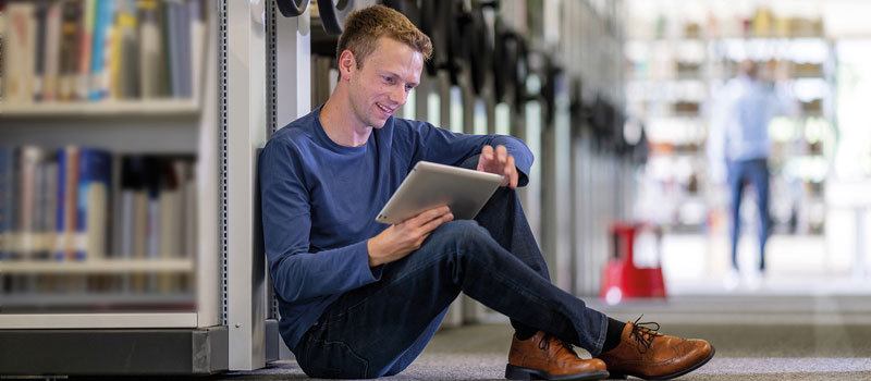 junger Mann sitzt in Bibliothek am Boden und arbeitet am Tablet