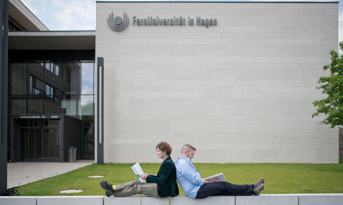 Zwei Personen sitzen Rücken and Rücken vor einem Gebäude der FernUniversität Hagen und lesen