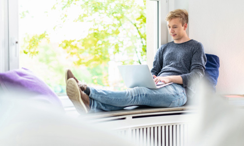 Junger Mann sitzt auf Fensterbank mit Laptop
