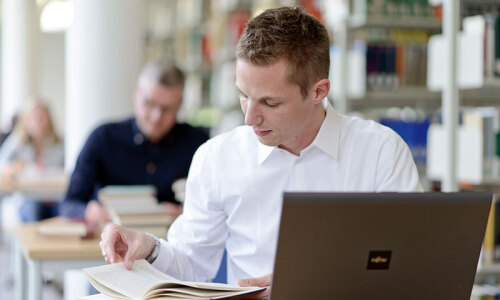 Ein Student sitzt in der Bibliothek vor einem Laptop und arbeitet mit einem Buch