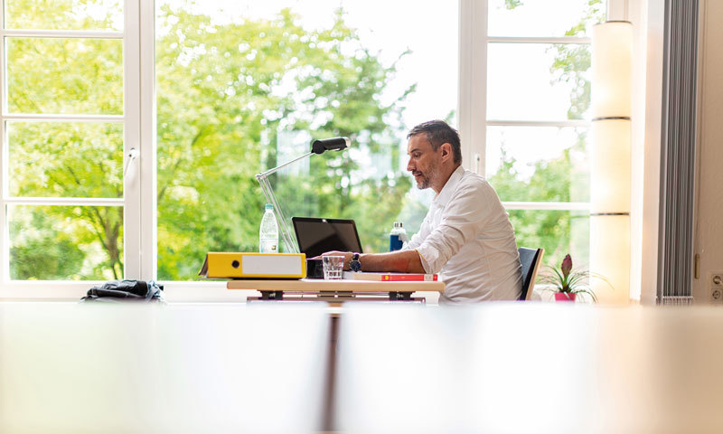Mann im mittleren Alter am Schreibtisch mit Laptop und einem gelben Ordner in einer häuslichen Umgebung vor einer offenen Fensterfront in grüner Umgebung 