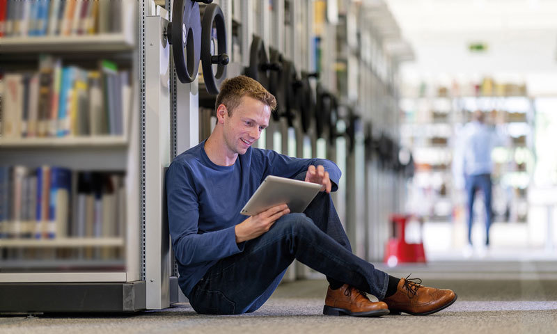 Ein junger Mann sitzt in einer Bibliothek auf dem Boden und arbeitet an einem Tablet.