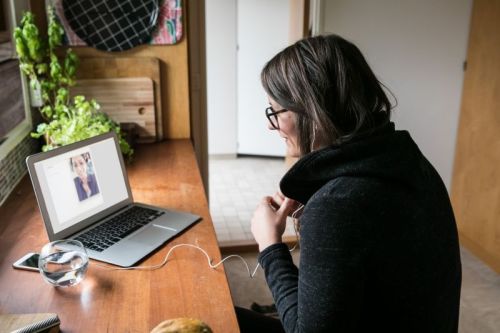 Eine Frau sitzt an einem schmalen Tisch und redet über ihren Laptop mit einer anderen Frau.