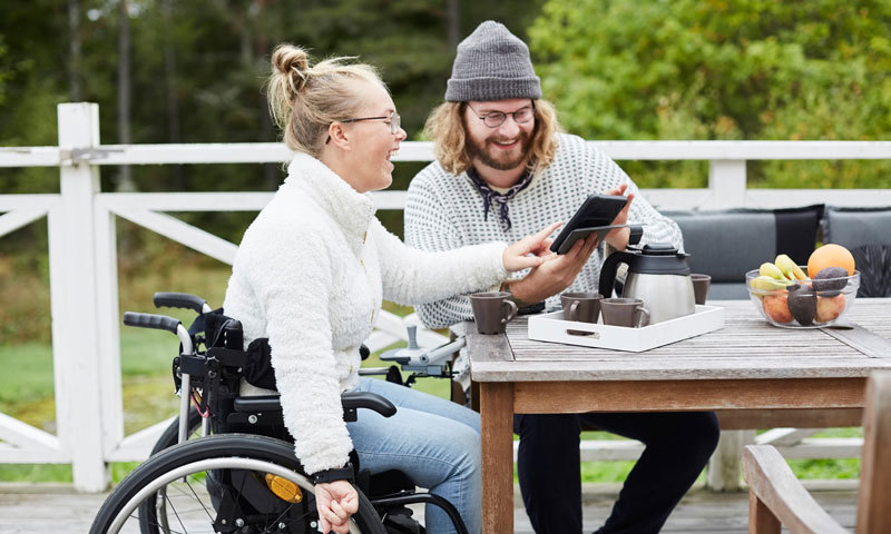Frau im Rollstuhl und junger Mann schauen gemeinsam auf ein Tablet und lachen