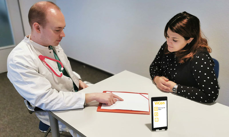 Ein Mann im Arztkittel und eine Frau sitzen an einem Tisch, auf dem ein Handy liegt mit dem Schriftzug ViCon auf dem Display.
