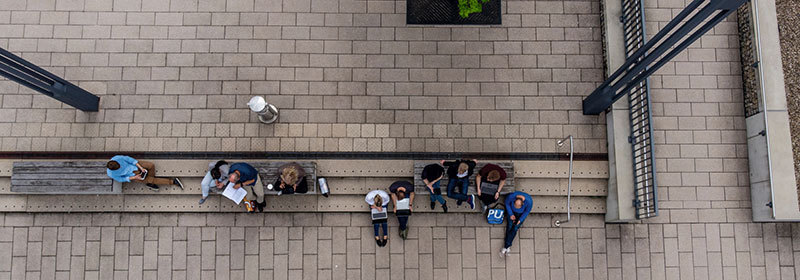 Campus aus der Vogelperspektive. Auf der Treppe sitzt eine Personengruppe, die gemeinsam auf ein Tablet schaut.