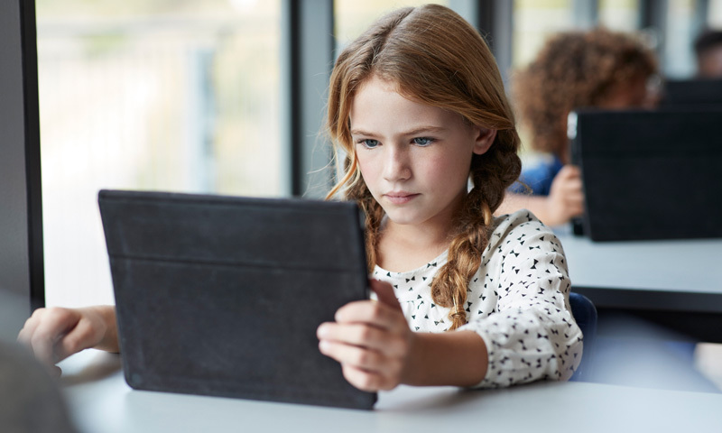 Mädchen hält Tablet in Klassenzimmer, schaut konzentriert