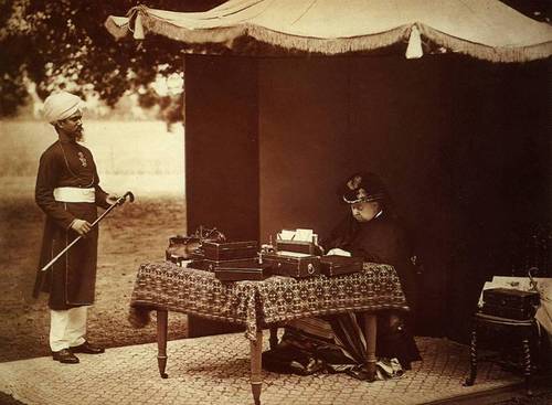 Foto in schwarz-weiß: Königin Victoria sitzt unter einem Baldachin im Freien an einer Schreibmaschine. Neben ihr steht ihr indischer Diener Abdul Karim.