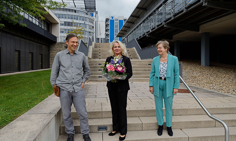 Drei lächelnde Personen auf Treppe, Frau in der Mitte mit Blumenstrauß