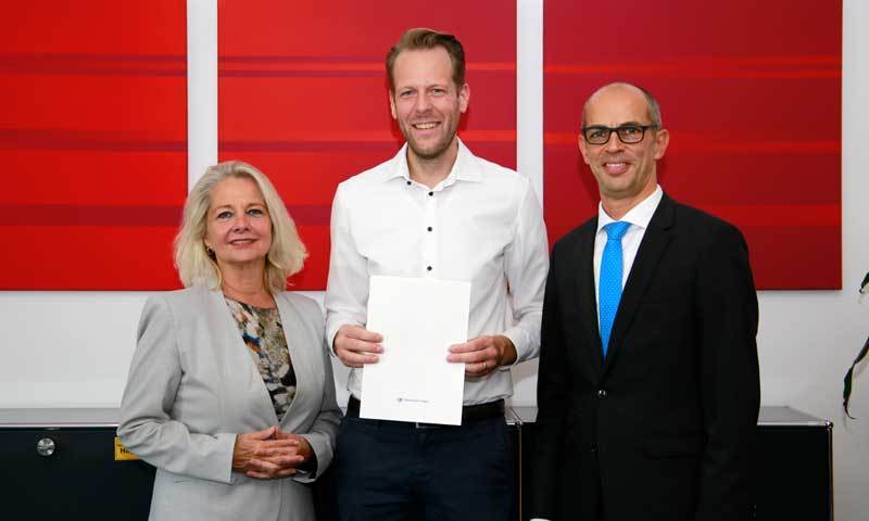 Juniorprofessor Michael Bucksteeg, FernUni-Rektorin Prof. Dr. Ada Pellert und Prof. Dr. Gerrit Brösel (Dekan der Fakultät für Wirtschaftswissenschaft) stehen vor roten Bildern in einem Büro. Michael Bucksteeg hält seine Ernennungsurkunde in der Hand.