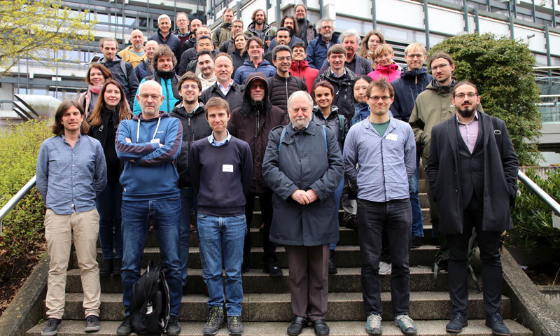 50 Forschende stehen für ein Gruppenbild auf einer Treppe. Im Hintergrund ist die FernUniversität zu sehen.