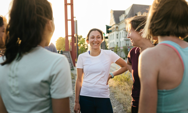 Gruppe von jungen sportlichen Frauen im Austausch. (Draußenaufnahme mit Häusern und rotem Metallmast im Hintergrund.)