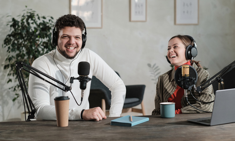 Männliche und weibliche Person bei einem Podcast mit Mikros und Headsets am Tisch mit aufgeschlagenem Laptop, blauem Notizblock und Kaffetassen vor einer Bilderwand.