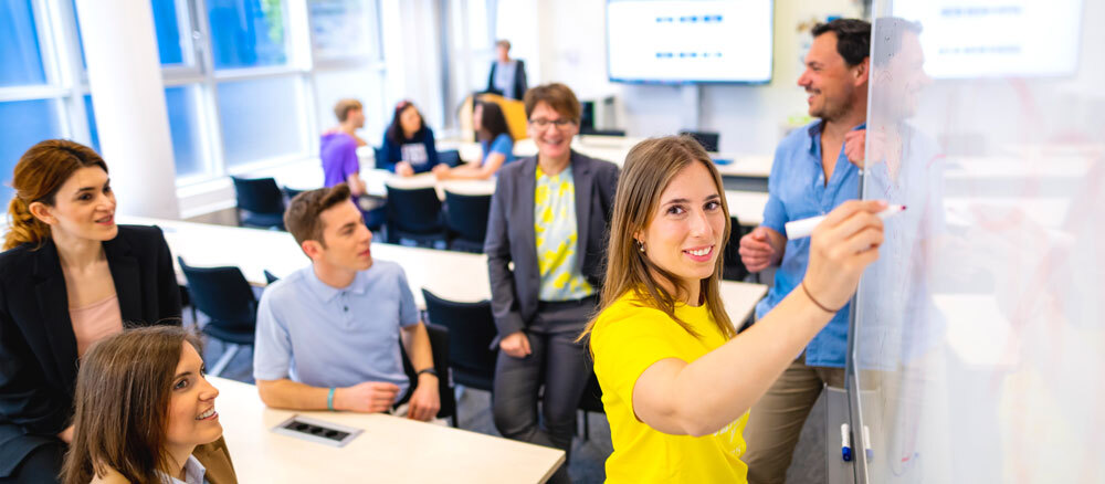 Eine Frau schreibt an ein Whiteboard. Sie befindet sich in einem Raum, in dem noch wietere Menschen stehen oder an Tischen sitzen.