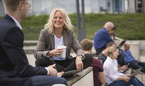 eine Frau mit Kaffeebecher sitzt auf dem Weißen Platz und unterhält sich mit einem Kommilitonen