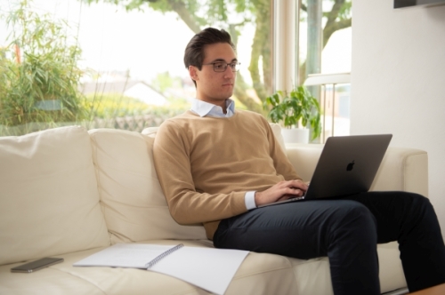 Ein junger Mann sitzt mit einem Laptop auf dem Schoß auf einem Sofa.