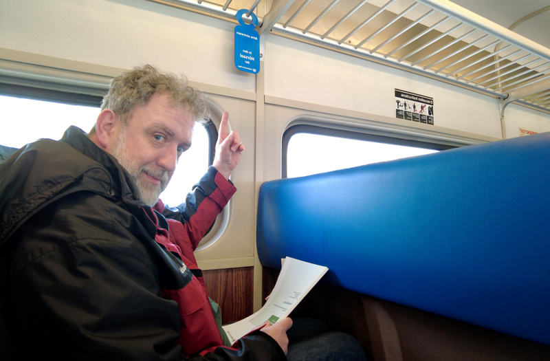 Ein Mann sitzt im Zugabteil und zeigt mit dem Finger auf ein Schild, das über ihm hängt, mit der Aufschrift Hörsaal der FernUni. 
