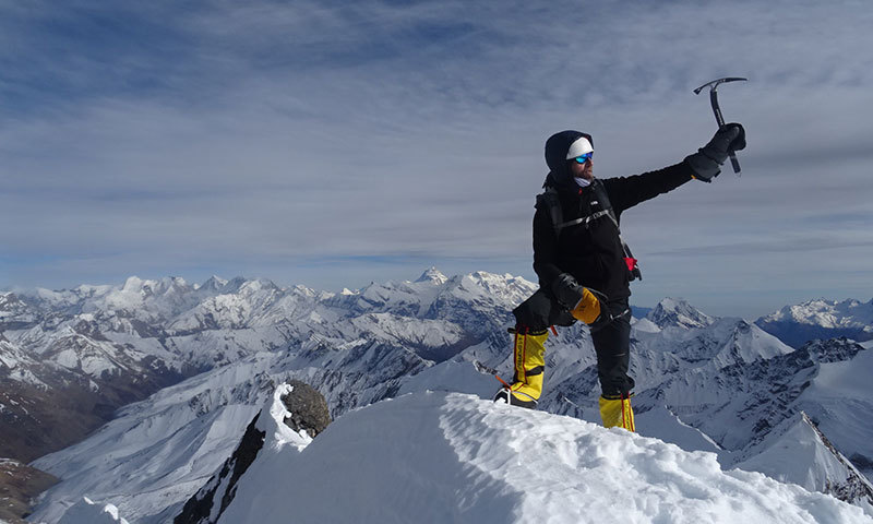 Mann mit Bergsteigerausrüstung steht auf einem verschneiten Berggipfel.
