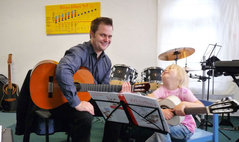 David Röhler sitzt mit einer Gitarre auf einem Stuhl in der Musikschule, neben ihm sitzt ein kleines Kind mit Gitarre.