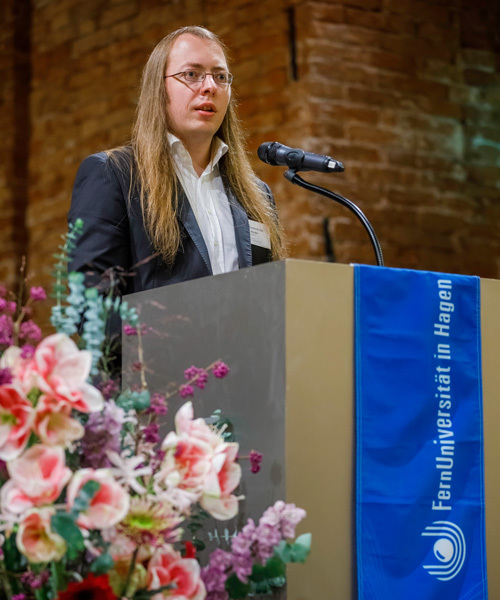 Mann steht am Rednerpult, Blumen im Vordergrund.