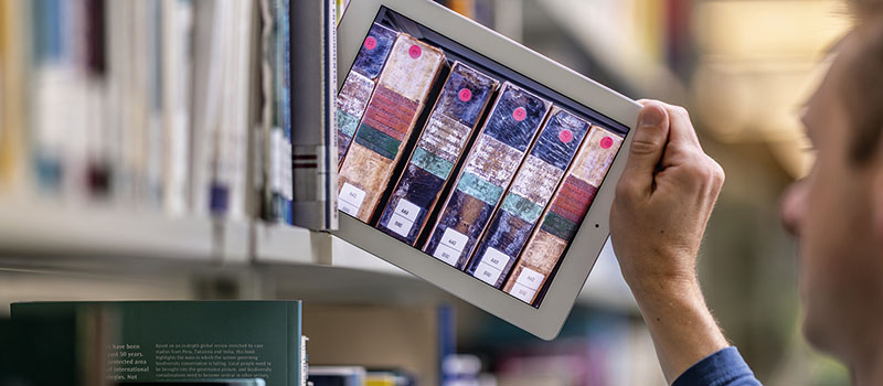 Tablet (statt Buch) wird aus einem Bücherregal rausgezogen. Auf dem Display sind alte Bücher (Rücken an Rücken) zu sehen.